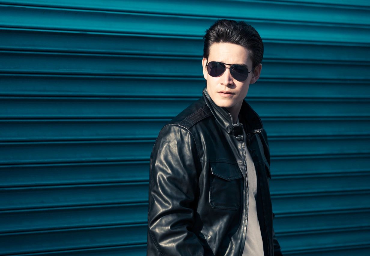 A man in sunglasses and leather jacket standing against a blue wall.